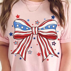 Puede incluir: Una camiseta rosa con un lazo rojo, blanco y azul con estrellas impresas.