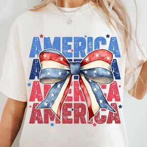 May include: Cream-colored t-shirt featuring a patriotic bow design with red, white, and blue stripes and stars. The word "AMERICA" is printed in blue and red, creating a layered effect. A casual and festive top.