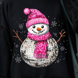May include: Black sweatshirt with a pink and silver snowman wearing a disco ball as a body. The snowman is wearing a pink hat and scarf with white snowflakes in the background.