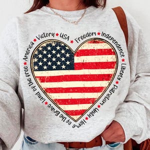 May include: Gray sweatshirt with a red, white, and blue heart-shaped American flag design. The heart is surrounded by a circle of text that reads "Home of the Brave Land of the Free America Victory USA Freedom Independence Liberty Patriotism Unity".