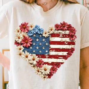 May include: Cream-colored t-shirt featuring a heart-shaped design of the American flag. The flag is made of distressed wood and surrounded by red, white, and blue flowers. The shirt has short sleeves and a crew neck.