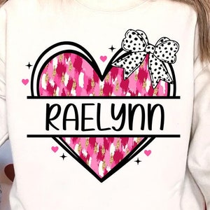 May include: A white sweatshirt with a pink and gold heart design and a black and white bow. The heart has the name "Raelynn" written across it.