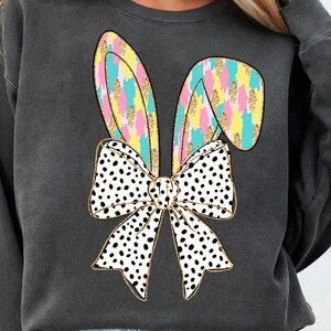 May include: A charcoal gray sweatshirt with a colorful bunny ear design and a black and white polka dot bow. The bow is tied around the bunny ears.
