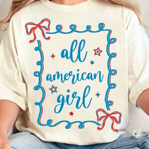 Peut inclure: Un t-shirt blanc avec un motif bleu et rouge qui dit "all american girl" &agrave; l'int&eacute;rieur d'un cadre bleu avec des &eacute;toiles rouges et des n&oelig;uds.