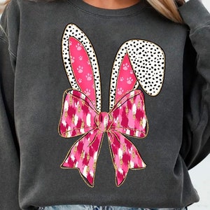 May include: A charcoal grey sweatshirt featuring a pink and white bunny ear design. The ears have a pink paw print pattern and the bow is pink with a white and gold glitter pattern.