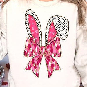 May include: A white sweatshirt featuring a pink and gold bow with bunny ears decorated with paw prints. The bow has a pink and white animal print.