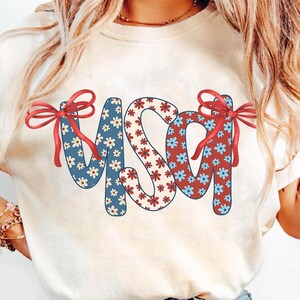 May include: A white t-shirt with a red, white, and blue floral design that spells out "USA" with red bows on each side of the letters.