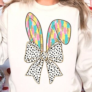 May include: A white sweatshirt with a colorful bunny ear design and a black and white polka dot bow. The bunny ears are pink, yellow, blue, and green with gold glitter accents.