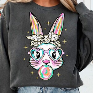May include: A dark gray sweatshirt with a white bunny wearing pink heart-shaped sunglasses and a black and white polka dot headband. The bunny is holding a pink and blue lollipop in its mouth.