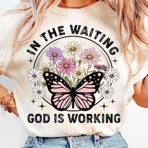 May include: A white t-shirt with a floral design and a butterfly. The text "In the waiting, God is working" is printed on the shirt.