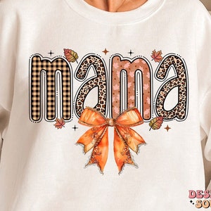 May include: White sweatshirt with the word "mama" in a fall-themed font with a plaid, leopard print, and leaf pattern. A large orange bow is below the word.