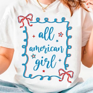 Peut inclure: T-shirt blanc avec un motif graphique bleu et rouge. Le graphique pr&eacute;sente un n&oelig;ud rouge et bleu, des &eacute;toiles et le texte "all american girl".