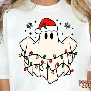 May include: White t-shirt with a cartoon ghost wearing a Santa hat and Christmas lights. The ghost has two black eyes and a red nose.