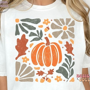 May include: White t-shirt featuring a graphic design with a pumpkin, leaves, acorns, and flowers in a retro style. The design is in shades of orange, brown, green, and beige.