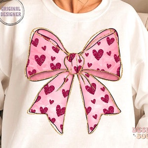 May include: A white sweatshirt with a pink and gold bow with a heart pattern. The bow is centered on the front of the sweatshirt.