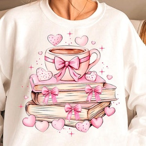 K&ouml;nnte beinhalten: Ein wei&szlig;er Pullover mit einer rosa und wei&szlig;en Illustration einer Tasse Kaffee, einem Stapel B&uuml;cher und Herzen.