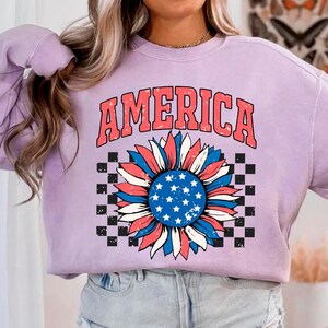 May include: A lavender sweatshirt with the word "AMERICA" printed in red, white, and blue. A red, white, and blue sunflower with a checkered background is printed below the word.