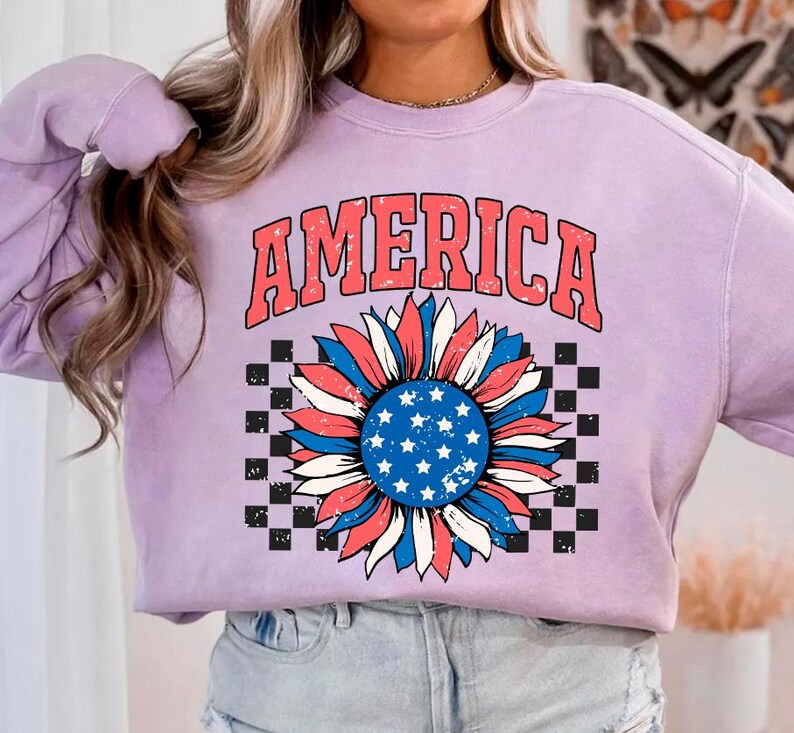 May include: A lavender sweatshirt with the word "AMERICA" printed in red, white, and blue. A red, white, and blue sunflower with a checkered background is printed below the word.