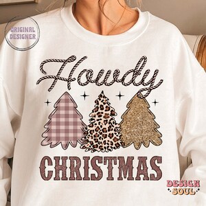 May include: White sweatshirt with the text "Howdy Christmas" in pink and brown. The text is surrounded by three Christmas trees, one with a pink and white plaid pattern, one with a leopard print, and one with gold glitter.