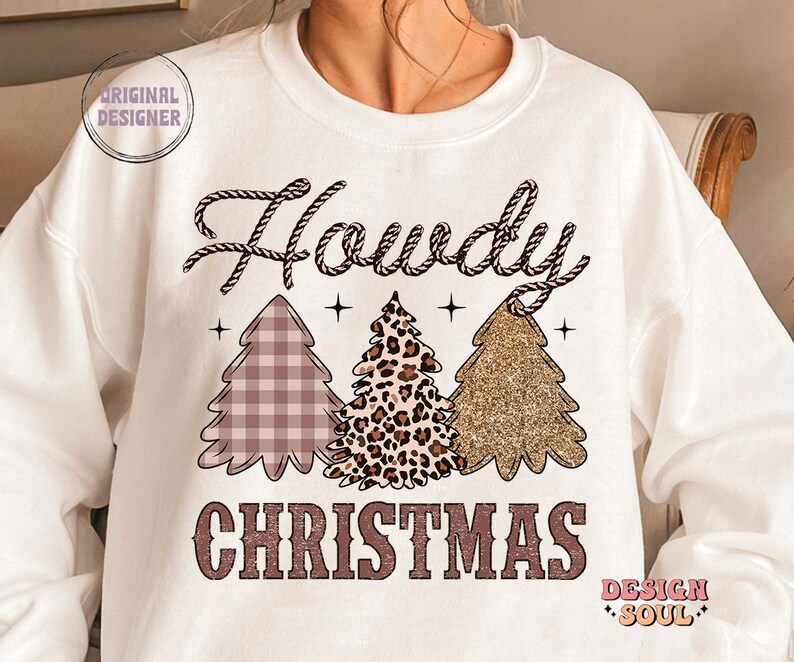 May include: White sweatshirt with the text "Howdy Christmas" in pink and brown. The text is surrounded by three Christmas trees, one with a pink and white plaid pattern, one with a leopard print, and one with gold glitter.