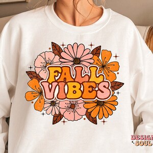 May include: White sweatshirt featuring a graphic design of orange, pink and brown flowers with the text "Fall Vibes" in a retro font.
