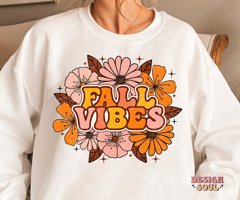 May include: White sweatshirt featuring a graphic design of orange, pink and brown flowers with the text "Fall Vibes" in a retro font.