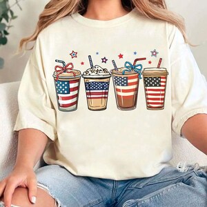 May include: A white t-shirt with a graphic of four iced coffee drinks decorated with the American flag. The coffee drinks are arranged in a row from left to right.
