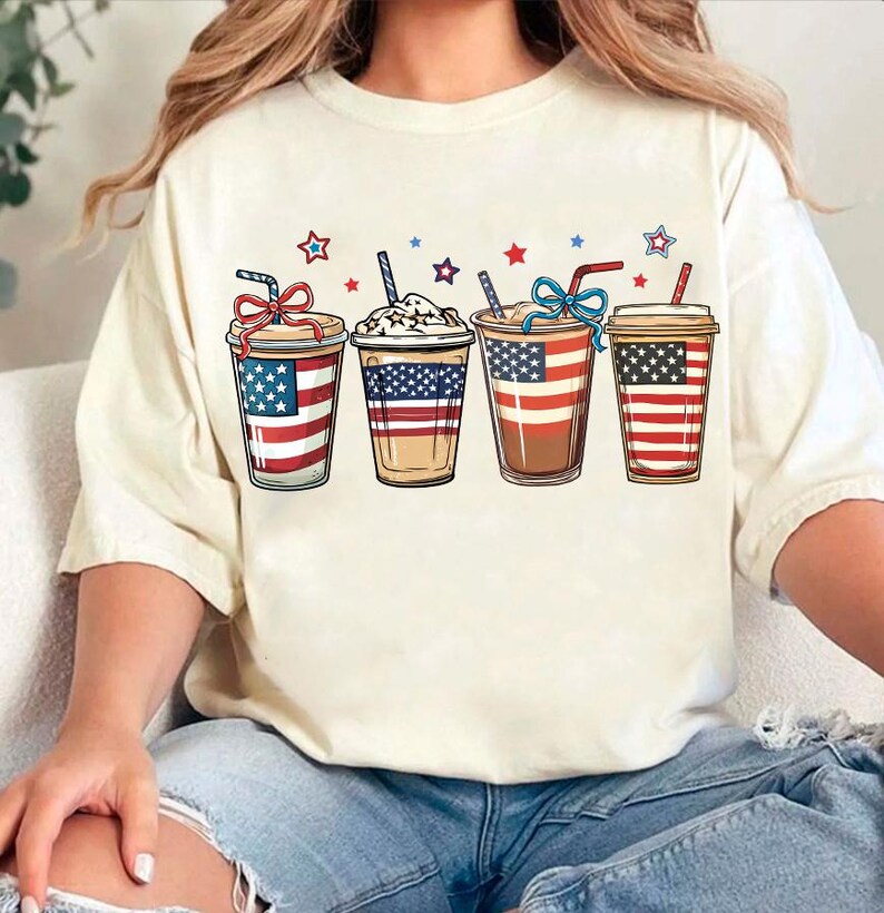 May include: A white t-shirt with a graphic of four iced coffee drinks decorated with the American flag. The coffee drinks are arranged in a row from left to right.