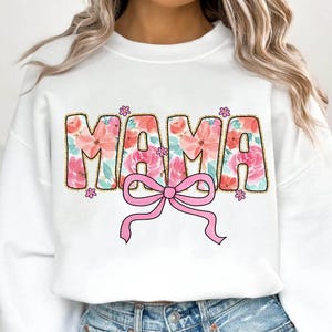 May include: White sweatshirt with the word "MAMA" in pink and gold letters with a floral pattern. The letters are outlined in gold and have a pink bow underneath.