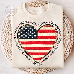 May include: A beige sweatshirt with a distressed American flag heart design. The heart is surrounded by the words "Victory", "USA", "Freedom", "Independence", "Liberty", "Patriotism", "Unity", "Home", "of the Brave", "Land of the free", and "America".