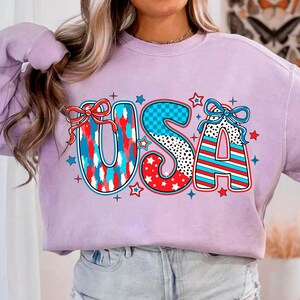 May include: A lavender sweatshirt with a graphic of the word "USA" in red, white, and blue with stars and checkered patterns.