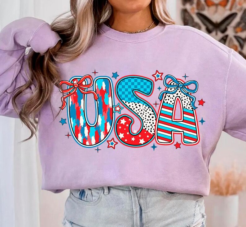 May include: A lavender sweatshirt with a graphic of the word "USA" in red, white, and blue with stars and checkered patterns.