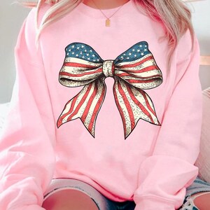 Peut inclure: Un sweat-shirt rose avec un grand n&oelig;ud rouge, blanc et bleu imprim&eacute; sur le devant. Le n&oelig;ud est dans le style du drapeau am&eacute;ricain.