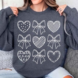 May include: A dark gray sweatshirt with a white design of hearts and bows. The hearts have different patterns, including polka dots, checkered, and stripes.