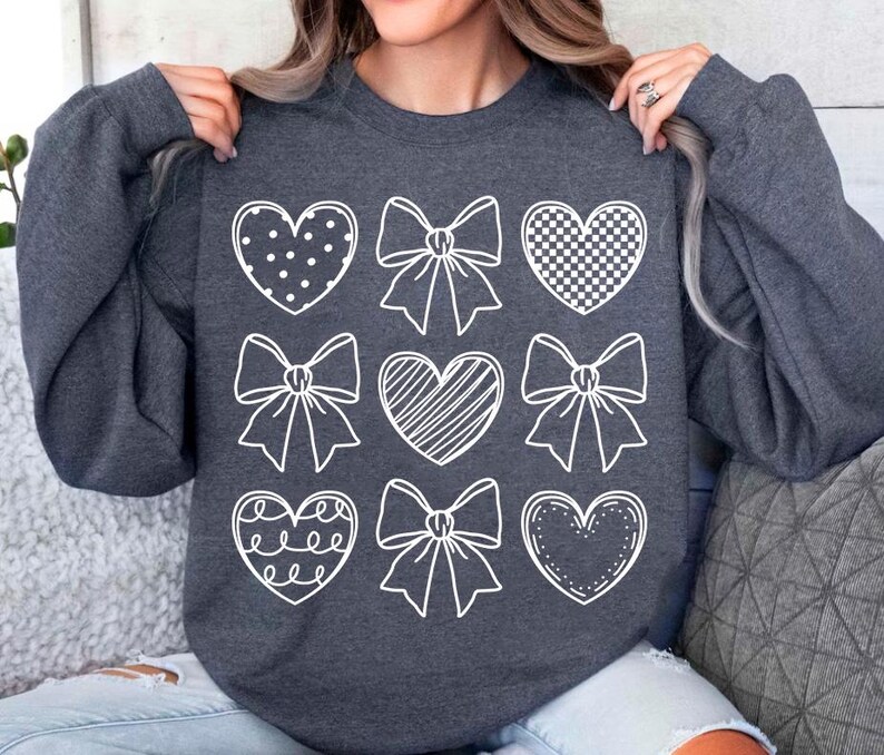 May include: A dark gray sweatshirt with a white design of hearts and bows. The hearts have different patterns, including polka dots, checkered, and stripes.