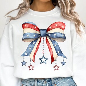 May include: White sweatshirt with a red, white, and blue bow with stars printed on it. The bow has strings with stars hanging from it.