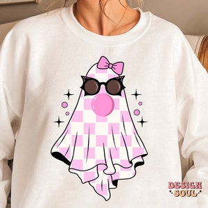 May include: White sweatshirt with a pink and white checkered ghost wearing sunglasses and a pink bow. The ghost has a pink bubble gum in its mouth. The text "DESIGN SOUL" is printed on the bottom right corner of the sweatshirt.