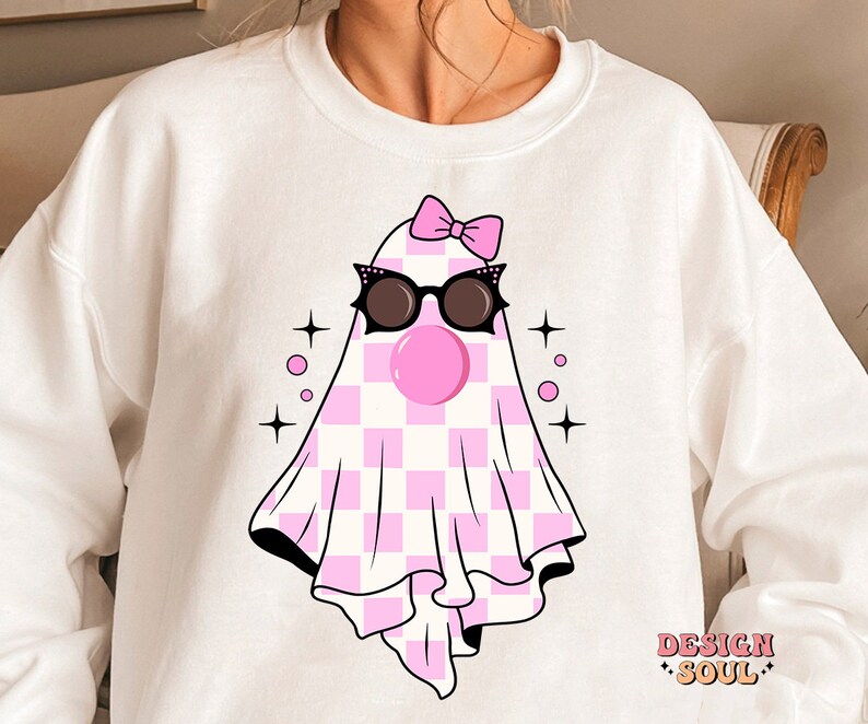 May include: White sweatshirt with a pink and white checkered ghost wearing sunglasses and a pink bow. The ghost has a pink bubble gum in its mouth. The text "DESIGN SOUL" is printed on the bottom right corner of the sweatshirt.