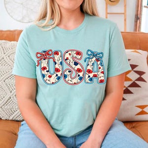 May include: A light blue t-shirt with the word "USA" printed on the front. The letters are outlined in red and blue and filled with a floral pattern featuring red and blue flowers.