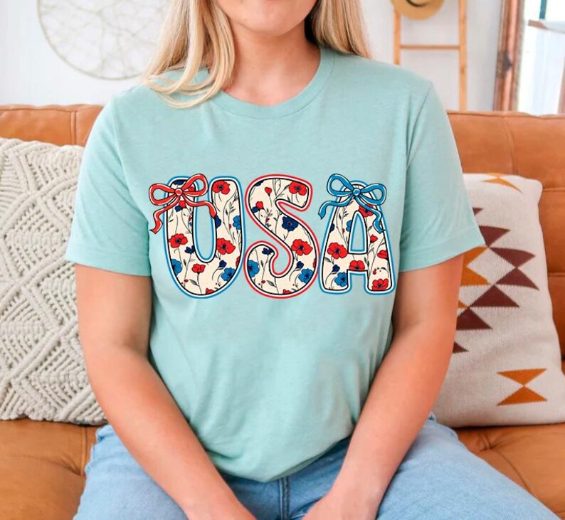 May include: A light blue t-shirt with the word "USA" printed on the front. The letters are outlined in red and blue and filled with a floral pattern featuring red and blue flowers.