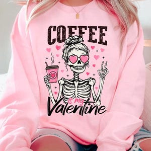 May include: Pink sweatshirt with a skeleton wearing heart-shaped sunglasses and holding a coffee cup. The text "COFFEE is my VALENTINE" is printed on the sweatshirt.