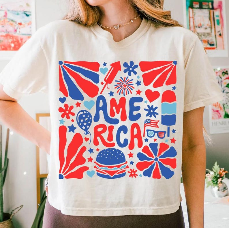 以下が含まれることがあります： 赤、白、青のグラフィックデザインが施された白いTシャツ。グラフィックにはレトロなフォントで「AMERICA」という言葉が書かれており、星、ハート、花火、その他の愛国的なシンボルで囲まれています。