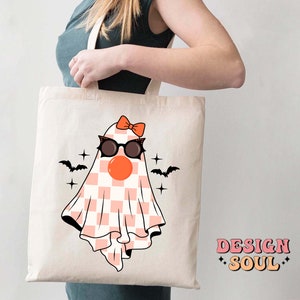 May include: A white tote bag with a pink and white checkered ghost wearing black sunglasses and a pink bow. The ghost has a red nose and there are black bats flying above it. The text "DESIGN SOUL" is printed in pink and orange on the bottom right corner of the bag.