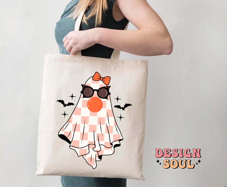 May include: A white tote bag with a pink and white checkered ghost wearing black sunglasses and a pink bow. The ghost has a red nose and there are black bats flying above it. The text "DESIGN SOUL" is printed in pink and orange on the bottom right corner of the bag.