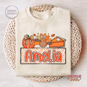 May include: A beige sweatshirt with a Thanksgiving-themed design featuring a pumpkin, a turkey, and a slice of pie. The design is surrounded by a leopard print border and the name "Amelia" is written in orange letters.