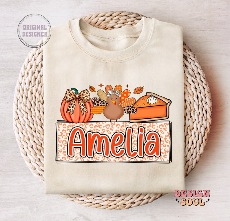 May include: A beige sweatshirt with a Thanksgiving-themed design featuring a pumpkin, a turkey, and a slice of pie. The design is surrounded by a leopard print border and the name "Amelia" is written in orange letters.