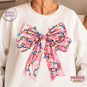 May include: A white sweatshirt with a pink bow decorated with colorful string lights. The bow is centered on the front of the sweatshirt. The text "DESIGN SOUL" is printed on the bottom right corner of the sweatshirt.