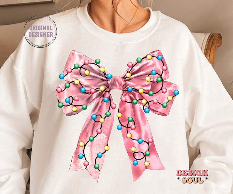 May include: A white sweatshirt with a pink bow decorated with colorful string lights. The bow is centered on the front of the sweatshirt. The text "DESIGN SOUL" is printed on the bottom right corner of the sweatshirt.