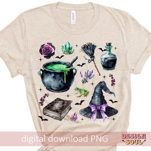 May include: A beige t-shirt with a watercolor design of a witch's cauldron, a witch's hat, a frog, a book, a bottle, a broom, and flowers. The design is in shades of green, purple, black, and brown.