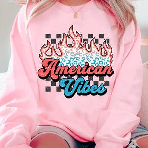 May include: Pink sweatshirt with a graphic of a flame with a leopard print design and the text "American Vibes" in a retro font.